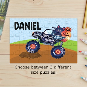 May include: A Halloween-themed puzzle featuring a black monster truck with spiderwebs, the text "Trick or Treat," and a black cat with pumpkins. The name "DANIEL" is printed in large black letters above the truck. The puzzle is on a light blue background.