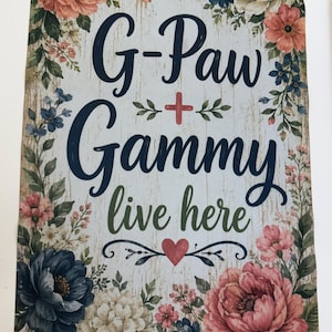 May include: A rectangular garden flag with a light wood-grain background featuring a floral border of pink, blue, and white flowers. The text reads G-Paw + Gammy live here in dark blue and green script, accented by a small red heart at the bottom.