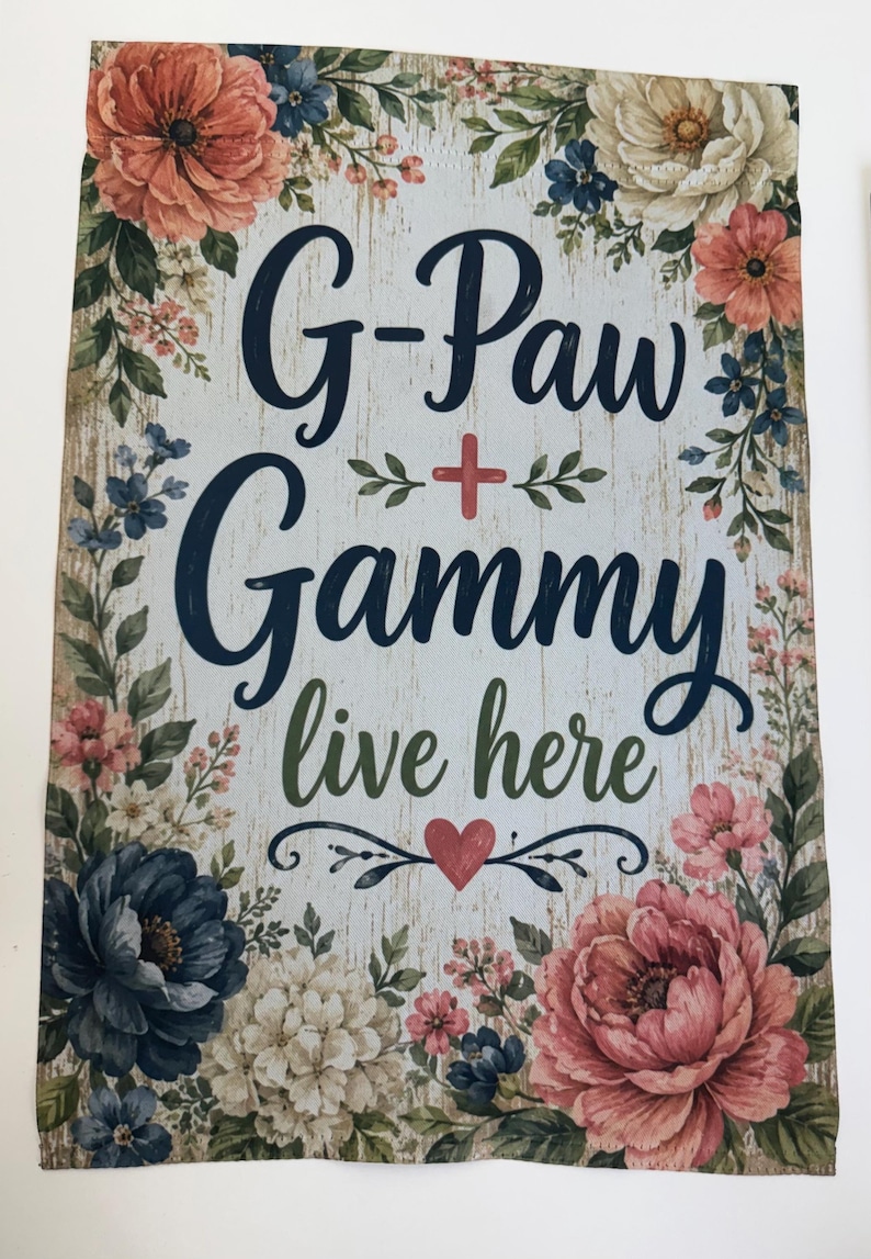 May include: A rectangular garden flag with a light wood-grain background featuring a floral border of pink, blue, and white flowers. The text reads G-Paw + Gammy live here in dark blue and green script, accented by a small red heart at the bottom.