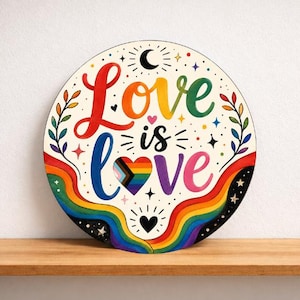 May include: A circular wall sign featuring the text Love is Love in colorful lettering. The design includes a rainbow-colored wave at the bottom, a crescent moon, stars, and a small heart with the Progress Pride flag pattern in the center.