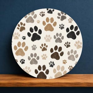 May include: A circular sign featuring a repeating pattern of animal paw prints in various shades of brown, gray, and black against an off-white background. The design includes multiple sizes of prints scattered across the entire surface of the disc.