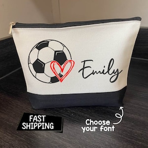 May include: A canvas cosmetic bag with a black base and zipper. The bag features a soccer ball graphic with a red heart and the name "Emily" in black script. The bag is white with a black trim.