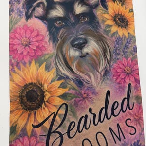 May include: A rectangular garden flag featuring a portrait of a black and grey Schnauzer dog surrounded by yellow sunflowers, pink zinnias, and purple lavender. The text "Bearded BLOOMS" is printed in black script and block lettering at the bottom.
