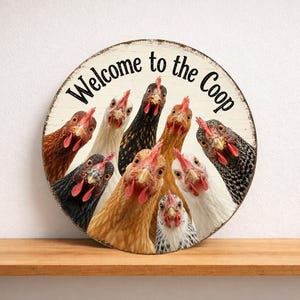 Può includere: Un cartello rotondo in legno con la scritta "Welcome to the Coop". Il cartello presenta un gruppo di galline con piume di vari colori, tra cui marrone, nero, bianco e beige, che sbucano dal bordo.