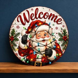 May include: A round sign featuring the word Welcome in red script at the top. The center shows a winking Santa Claus wearing a red suit and black belt, tangled in colorful glowing Christmas lights. Green holly leaves and red berries decorate the sides.