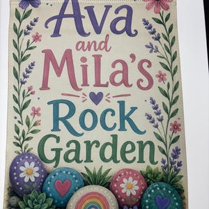 May include: A rectangular garden flag featuring the text Ava and Mila's Rock Garden in purple, pink, blue, and green lettering. The design is framed by floral vines and includes illustrations of painted rocks with daisies, hearts, and a rainbow.
