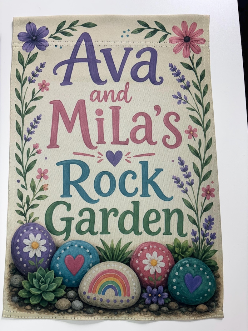 May include: A rectangular garden flag featuring the text Ava and Mila's Rock Garden in purple, pink, blue, and green lettering. The design is framed by floral vines and includes illustrations of painted rocks with daisies, hearts, and a rainbow.