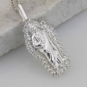May include: A silver pendant necklace featuring a detailed religious figure surrounded by a halo of sparkling cubic zirconia stones.