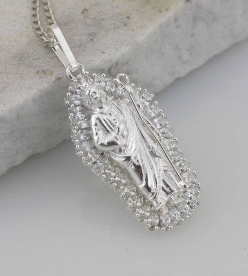 May include: A silver pendant necklace featuring a detailed religious figure surrounded by a halo of sparkling cubic zirconia stones.