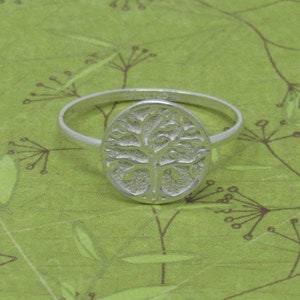 May include: A silver ring with a tree of life design. The ring is on a green background.