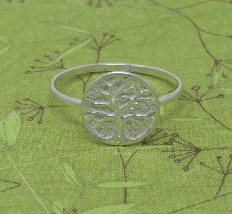 May include: A silver ring with a tree of life design. The ring is on a green background.