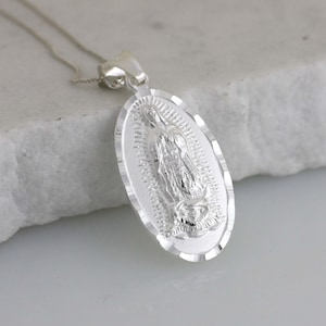 Pode incluir: Um colar com pingente oval prateado com uma representação detalhada de uma figura religiosa. O pingente tem uma borda texturizada e é suspenso por uma corrente delicada. O design é muito detalhado e o pingente é reflexivo.