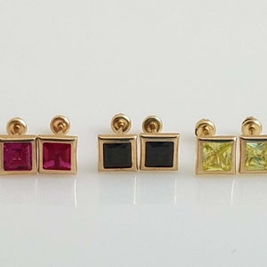 Op de afbeelding: Zes paar gouden vierkante oorbel studs met verschillende gekleurde edelstenen. De oorbellen hebben een vierkante zetting met een kleine edelsteen in het midden. De edelstenen zijn helder, rood, zwart, geel en paars.