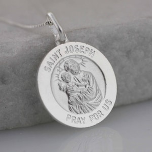 May include: Silver pendant necklace featuring a round charm with a depiction of Saint Joseph holding baby Jesus. The charm is inscribed with "Saint Joseph Pray For Us".