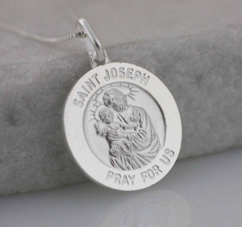 May include: Silver pendant necklace featuring a round charm with a depiction of Saint Joseph holding baby Jesus. The charm is inscribed with "Saint Joseph Pray For Us".
