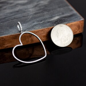 Op de afbeelding: Een zilveren hartvormige hanger met een rand bezet met diamanten. De hanger wordt getoond naast een Amerikaanse kwart voor groottevergelijking.