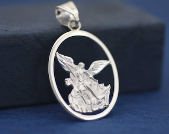 May include: A silver pendant featuring an angel within an oval frame. The angel has outstretched wings and is depicted in a detailed, sculpted style. The pendant has a loop at the top for a chain and is set against a dark blue background.