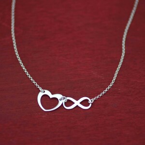 May include: A silver necklace with a heart and infinity symbol pendant. The necklace is on a red background.