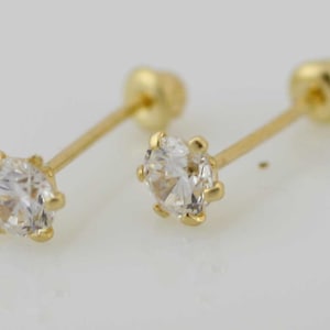 May include: Two gold stud earrings with clear, round cubic zirconia stones. The earrings have a post and screw-back closure. The gold setting has a six-prong design, holding the stone securely. The earrings are displayed against a light background.