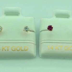 May include: Two pairs of stud earrings on white display cards. One pair features clear gemstones, the other pair has red gemstones. The cards are labeled "14 KT GOLD" in gold lettering.