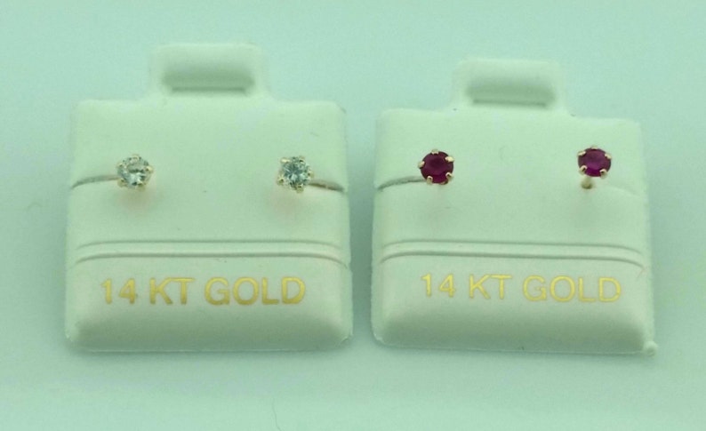 May include: Two pairs of stud earrings on white display cards. One pair features clear gemstones, the other pair has red gemstones. The cards are labeled "14 KT GOLD" in gold lettering.