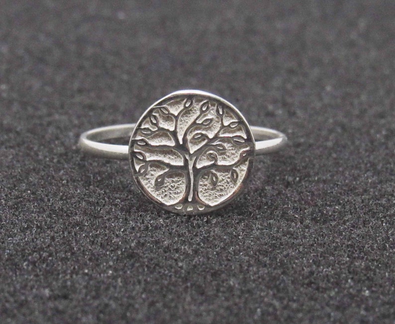 May include: A silver ring with a tree of life design. The ring is round and has a detailed design of a tree with branches and leaves.