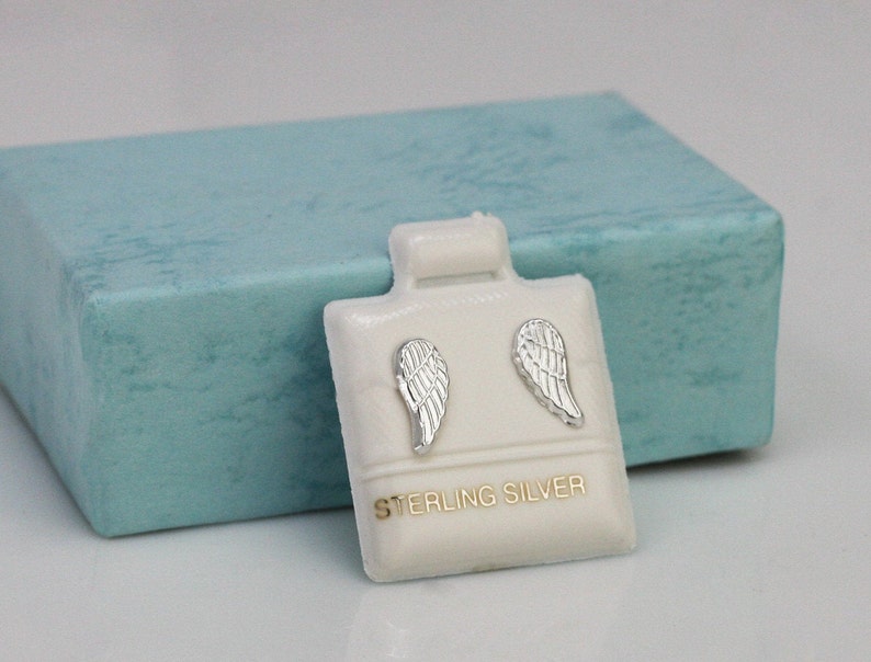 K&ouml;nnte beinhalten: Ein Paar Ohrringe aus Sterlingsilber in Form von Engelsfl&uuml;geln in einer wei&szlig;en Schachtel mit blauem Deckel. Die Ohrringe sind klein und zart und w&auml;ren ein tolles Geschenk f&uuml;r einen geliebten Menschen.
