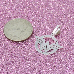 May include: Silver charm pendant featuring the Wonder Woman logo. The logo is a stylized 'W' with wings and stars around the circle. The pendant is on a pink glitter background.