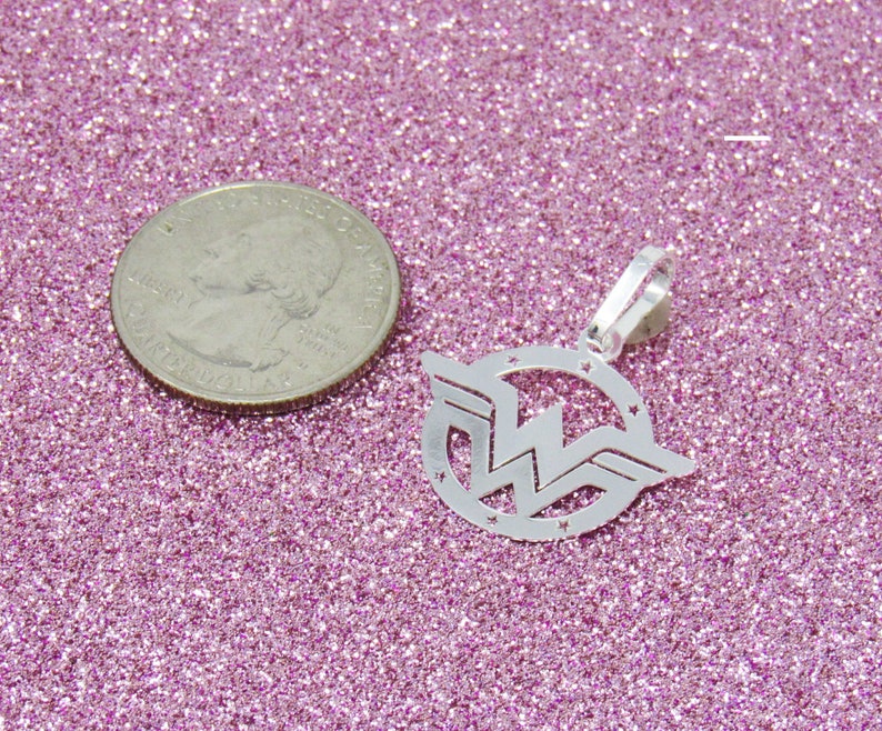 May include: Silver charm pendant featuring the Wonder Woman logo. The logo is a stylized 'W' with wings and stars around the circle. The pendant is on a pink glitter background.
