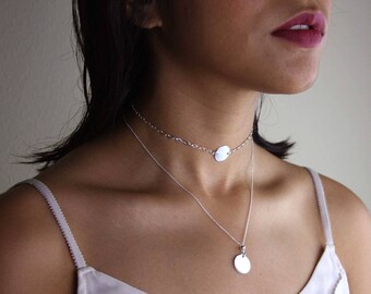 May include: A silver chain necklace with two circular pendants. The necklace is layered, with the top chain being a choker style and the bottom chain extending to the chest. The pendants are both plain and smooth.
