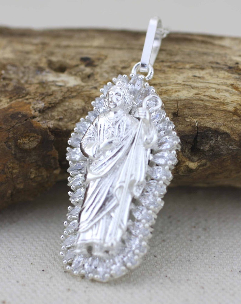 May include: Silver pendant featuring a detailed depiction of a religious figure with a staff, surrounded by a halo of sparkling stones.