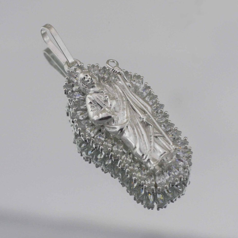 May include: Silver pendant featuring a religious figure surrounded by a halo of clear cubic zirconia stones.