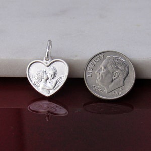 May include: Silver heart-shaped pendant featuring a detailed image of two angels embracing. The pendant is shown next to a US dime for size comparison.