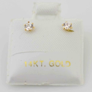 May include: A pair of gold stud earrings with clear, round gemstones. The earrings are set in a six-prong setting and are displayed on a white card with the text "14KT. GOLD" in gold lettering.