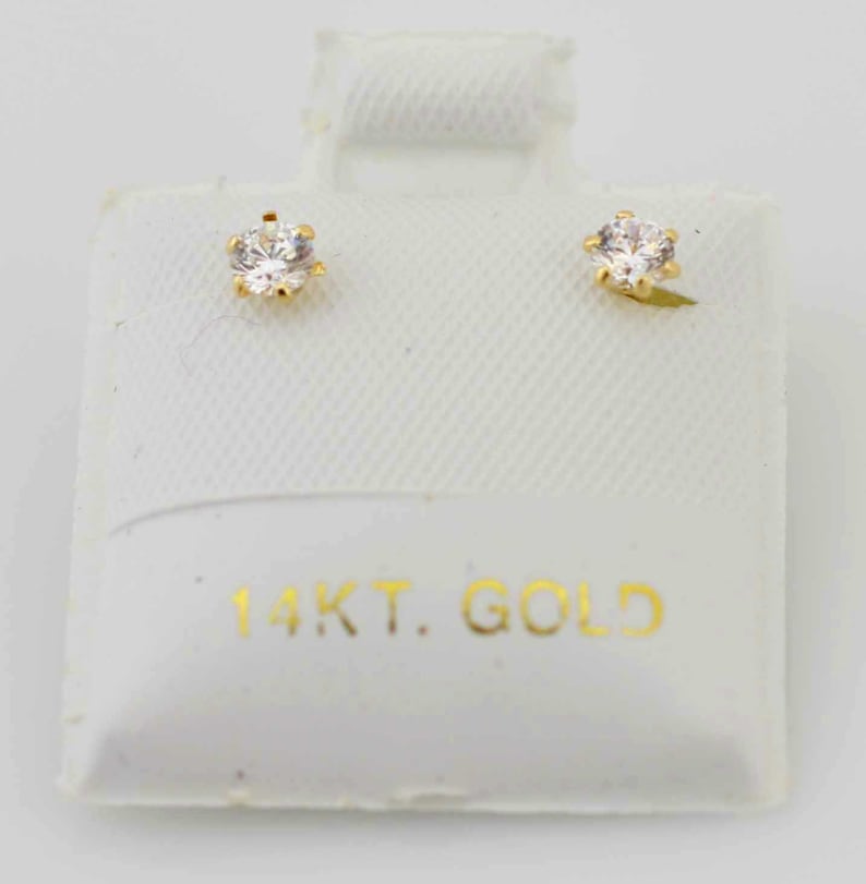 May include: A pair of gold stud earrings with clear, round gemstones. The earrings are set in a six-prong setting and are displayed on a white card with the text "14KT. GOLD" in gold lettering.