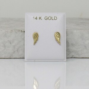 Puede incluir: Un par de pendientes de oro en forma de alas de &aacute;ngel en una tarjeta de presentaci&oacute;n blanca con el texto "14K GOLD".