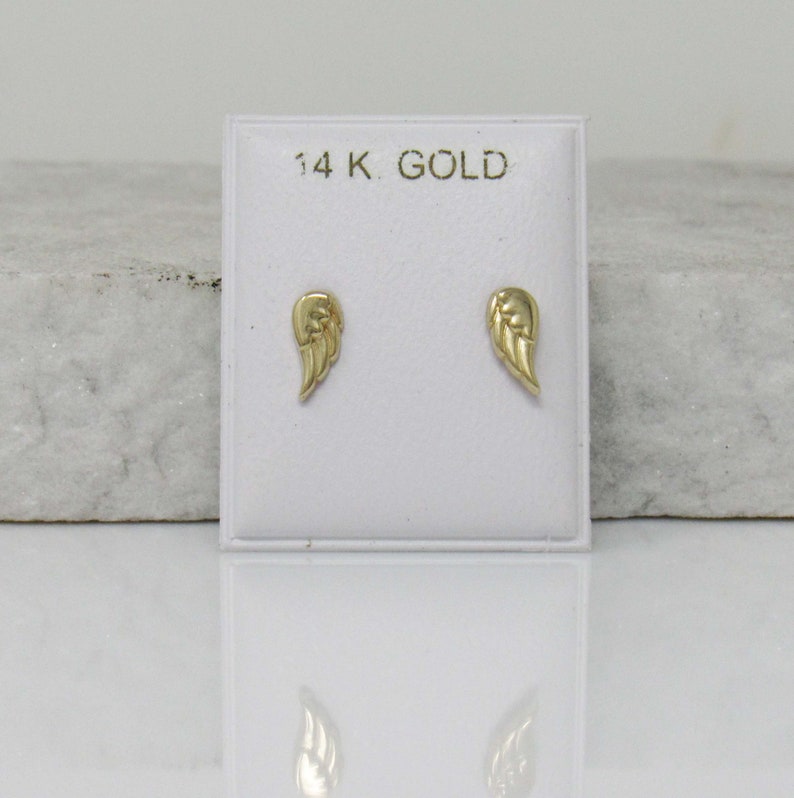 Puede incluir: Un par de pendientes de oro en forma de alas de &aacute;ngel en una tarjeta de presentaci&oacute;n blanca con el texto "14K GOLD".