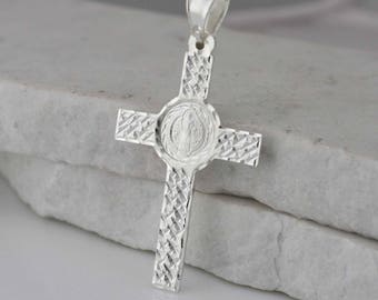 May include: Silver pendant featuring a cross with a detailed, textured design. The center of the cross has a round medallion with a depiction of Saint Benedict.