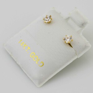 May include: A pair of gold stud earrings with clear, faceted gemstones. The earrings are displayed on a white card with the text "14KT. GOLD" printed in gold. The earrings have a classic, elegant design.