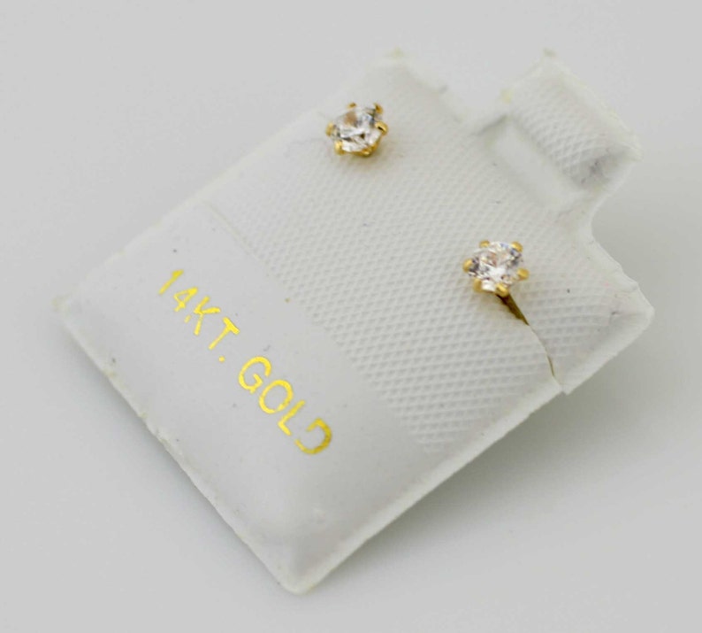 May include: A pair of gold stud earrings with clear, faceted gemstones. The earrings are displayed on a white card with the text "14KT. GOLD" printed in gold. The earrings have a classic, elegant design.
