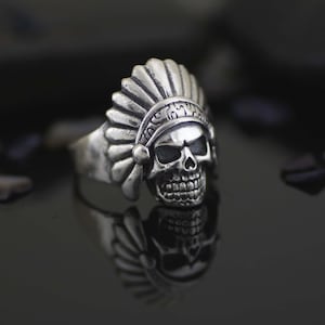 May include: A silver ring featuring a detailed skull design with a headdress resembling a Native American headdress.