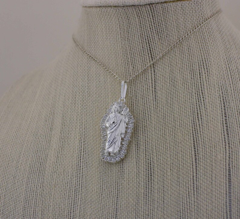 May include: A silver chain necklace with a pendant featuring a detailed image of Jesus Christ. The pendant is surrounded by a decorative border.