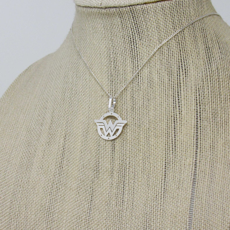 May include: A silver necklace with a Wonder Woman logo pendant. The pendant is a circular cutout of the logo with a detailed design.