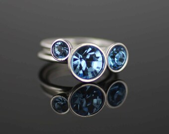 May include: A silver ring with three blue gemstones of varying sizes. The largest gemstone is centered, with two smaller ones flanking it. The ring is set against a dark, reflective surface, creating a mirrored effect.
