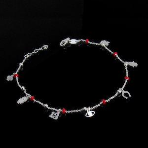 May include: Silver anklet with red beads and charms. Charms include a four-leaf clover, a teddy bear, the number 13, an eye, a horseshoe, and a hand. The anklet is on a black background.