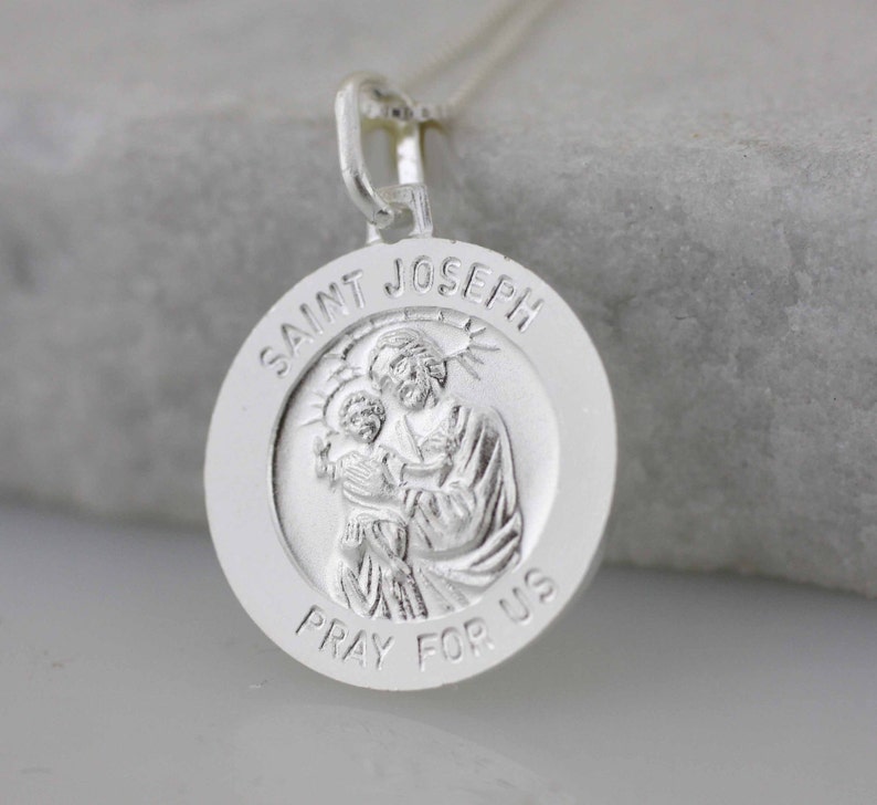 May include: Silver pendant necklace featuring a round medal with a depiction of Saint Joseph holding baby Jesus. The medal is inscribed with "Saint Joseph Pray For Us".