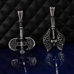 May include: Two silver-toned pendants with intricate designs. One pendant is an axe shape with a detailed blade and handle. The other pendant has a symmetrical, ornate design. Both pendants have a loop for a chain.