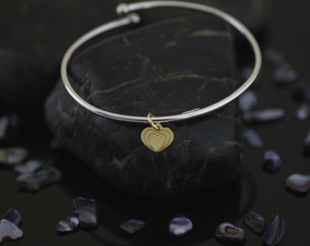 Puede incluir: Un brazalete de plata con un dije de corazón de oro. El dije está hecho de dos corazones superpuestos.