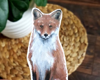 May include: A hand-painted sticker of a red fox sitting with its tail curled behind it. The fox has a white chest and a brown nose.