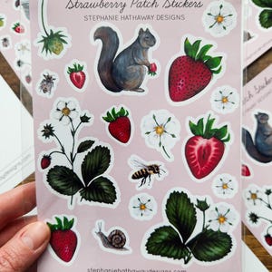Op de afbeelding: Een vel met stickers met verschillende illustraties met betrekking tot aardbeien, waaronder een eekhoorn, een bij, een slak en aardbeiplanten met bloemen en bladeren. De stickers zijn op een roze achtergrond en hebben de tekst "Strawberry Patch Stickers" en "Stephanie Hathaway Designs" erop gedrukt.